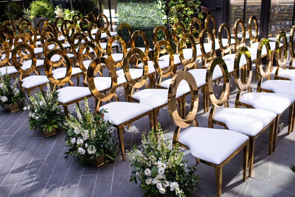Gold wedding ceremony chairs lined with white and greenery floral arrangements