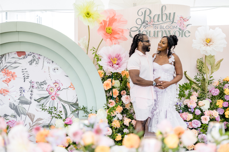 Mother-to-be and father pose in front of custom signage at their colorful baby shower.