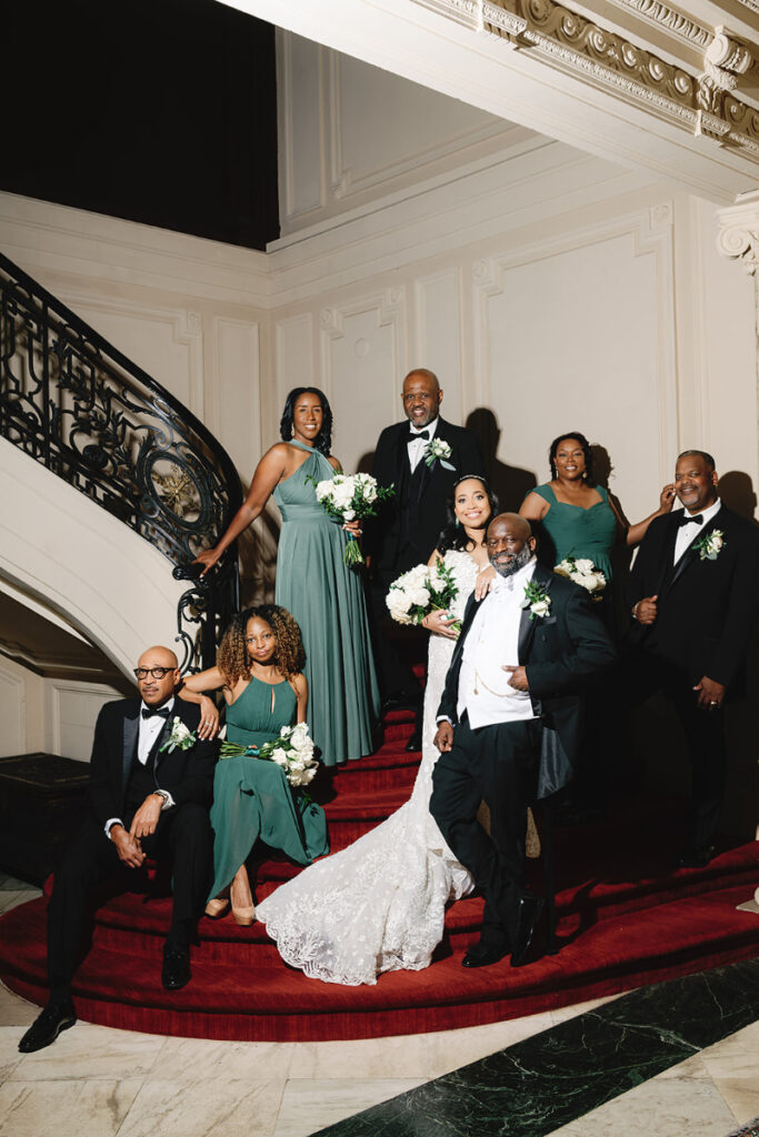 Bride and groom pose with wedding party on red carpet staircase.
