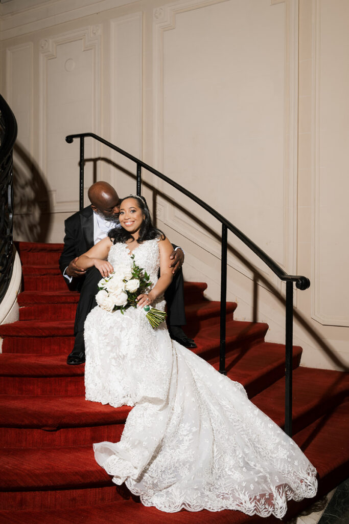 Bride and groom sit for photo on red carpet stairs.