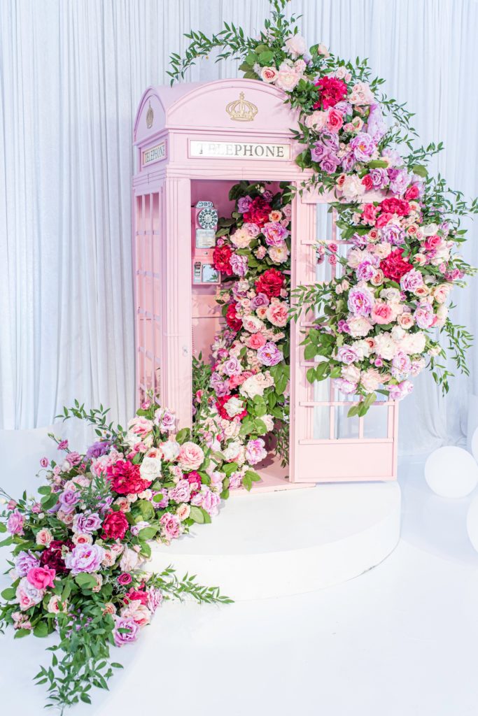 Luxury wedding shower photobooth backdrop using a pink telephone booth that is covered from top to bottom in a luxe stand of pink and purple flowers.