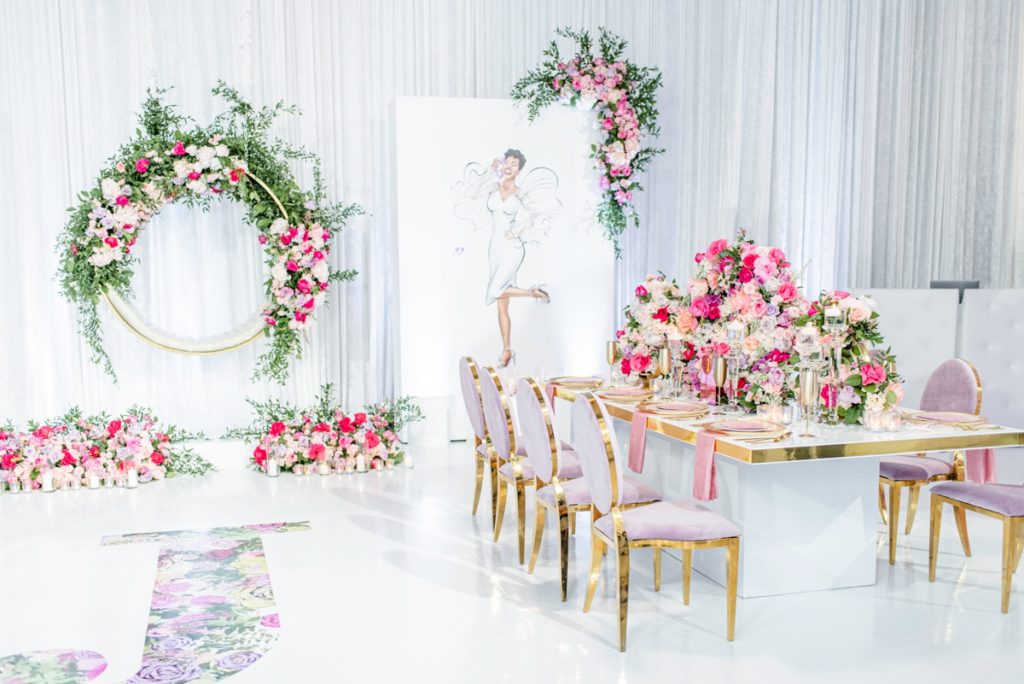 Luxe pink bridal shower decor with a personalized floral monogram floor decal, a circular flower swing photo backdrop, sketch of the bride to be, and a floral centerpieces with gold and pink table settings.