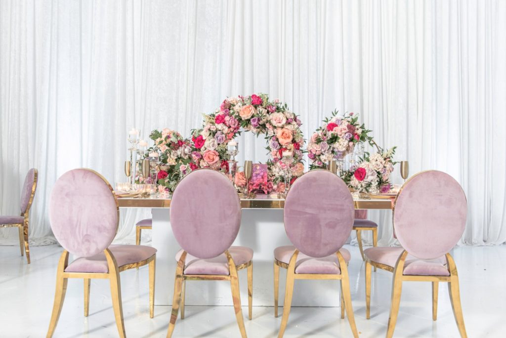 Luxury bridal shower decor with a gold table, pink velvet and gold chairs, gold champagne flutes and circular floral centerpieces on a white drapery backdrop
