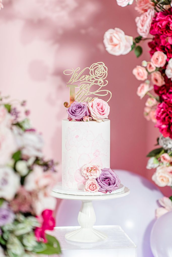 White bridal shower cake decorated with pink watercolor flowers, fresh flowers, and a gold cake topper.