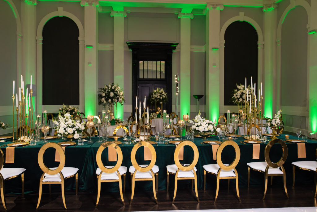 Long wedding reception table with emerald green linen, tall candles, and gold chairs