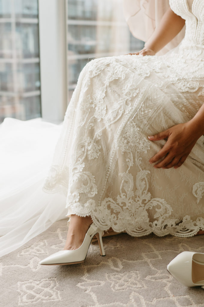 The bride's holding her lace dress while putting on her wedding heels