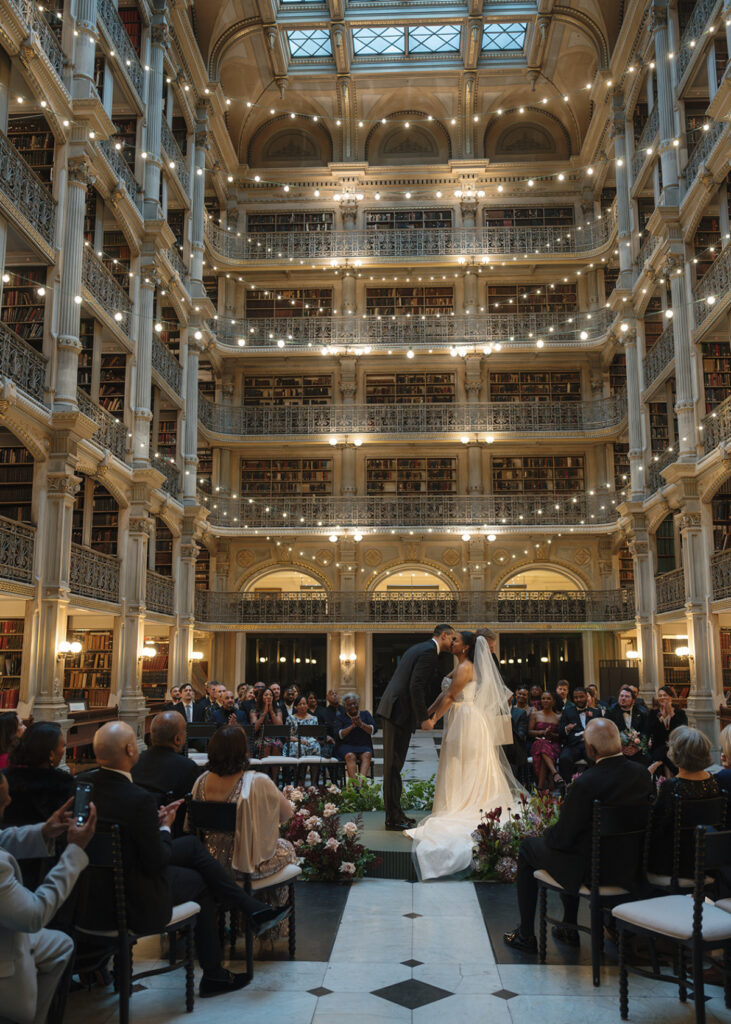 Bride and groom share their first kiss at a historic library wedding venue beneath the George Peabody Library's grand architecture.
