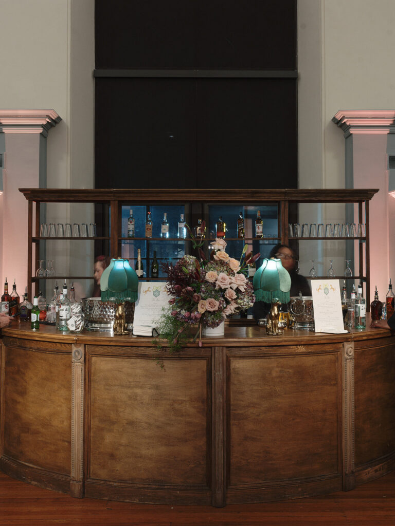 Vintage wood bar with custom cocktail menus, floral arrangement, and fringed lamps at the wedding reception.