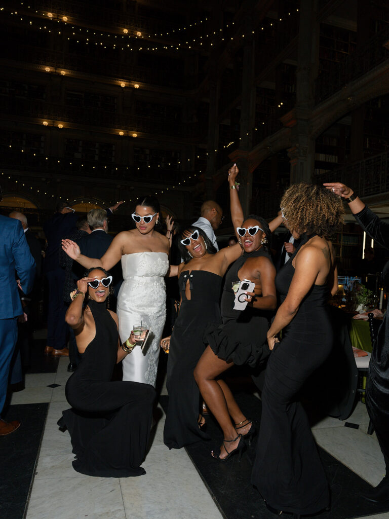 Bride and bridesmaids celebrate on the dance floor wearing fun heart-shaped sunglasses during the wedding reception.