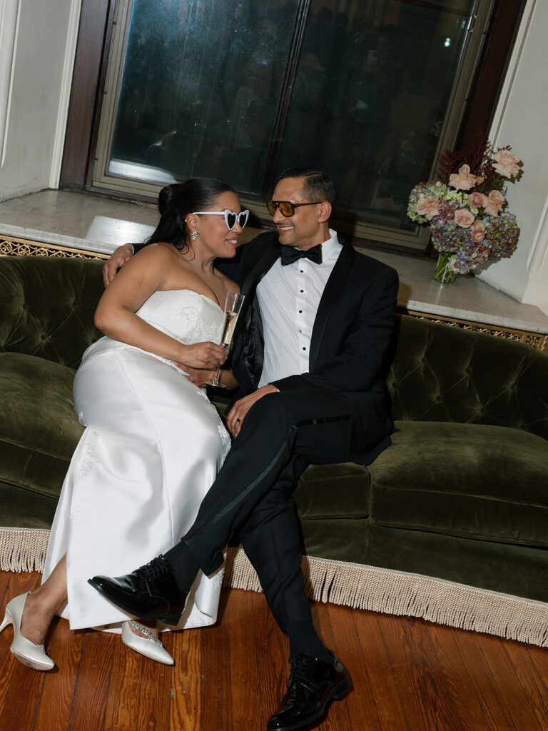 Bride and groom relax on a velvet sofa wearing fun sunglasses and sharing a champagne toast at the reception.
